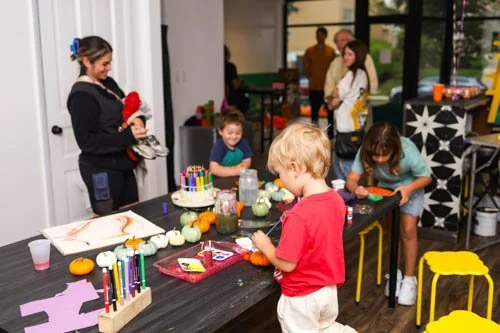 Kids and adults gathered around a table with Halloween decorations, candles, and pumpkin crafts, celebrating in a decorated room.