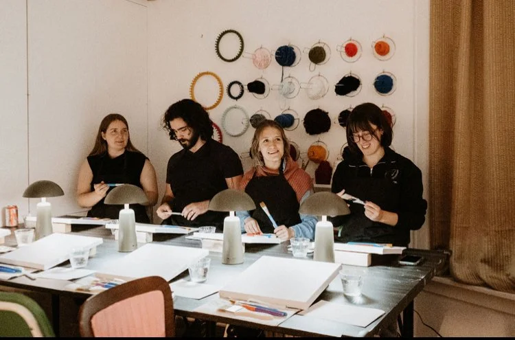 Four people seated at a long table with art supplies, in front of a wall decorated with colorful hoop earrings all hanging on hooks.