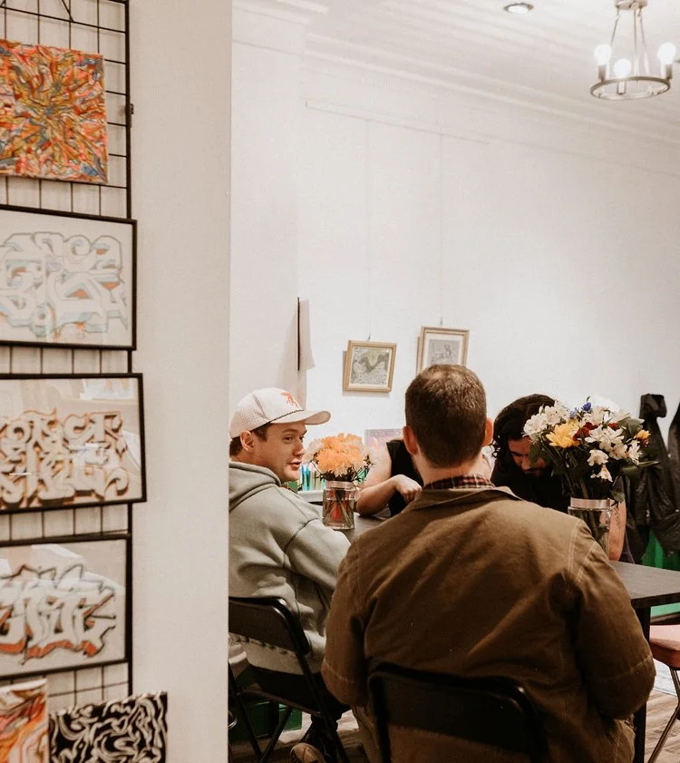 People sitting at a table with flower arrangements, talking in an art gallery or cafe setting with framed artwork on the walls.