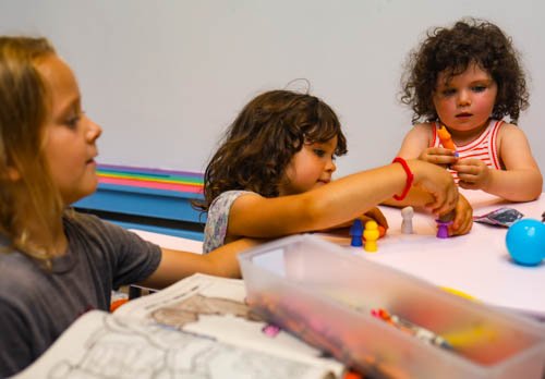 Three children sitting at a table playing with small toys, with a woman watching nearby.