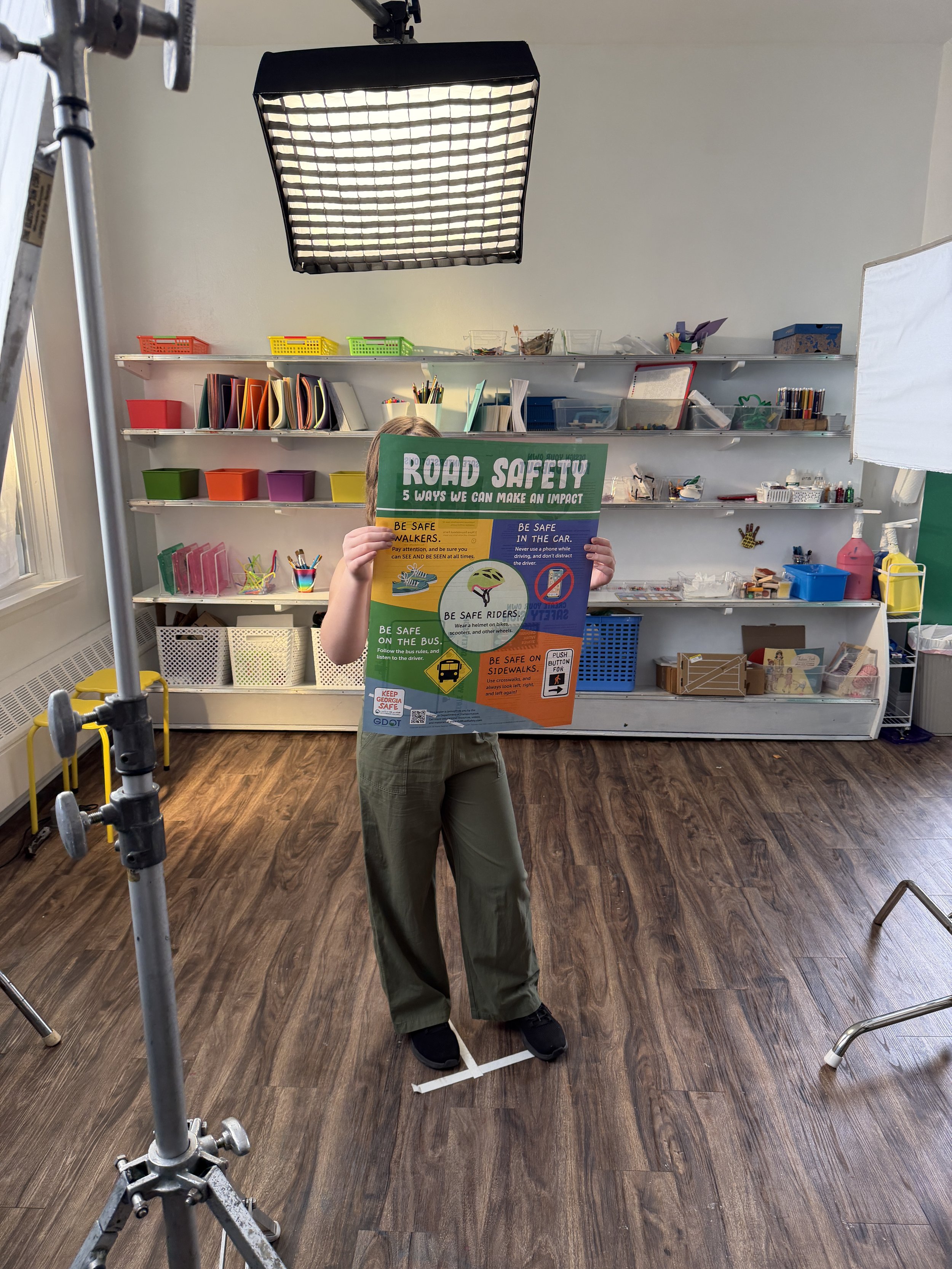 A person holding a colorful road safety poster in a room with wooden flooring and shelves filled with school supplies and art materials.
