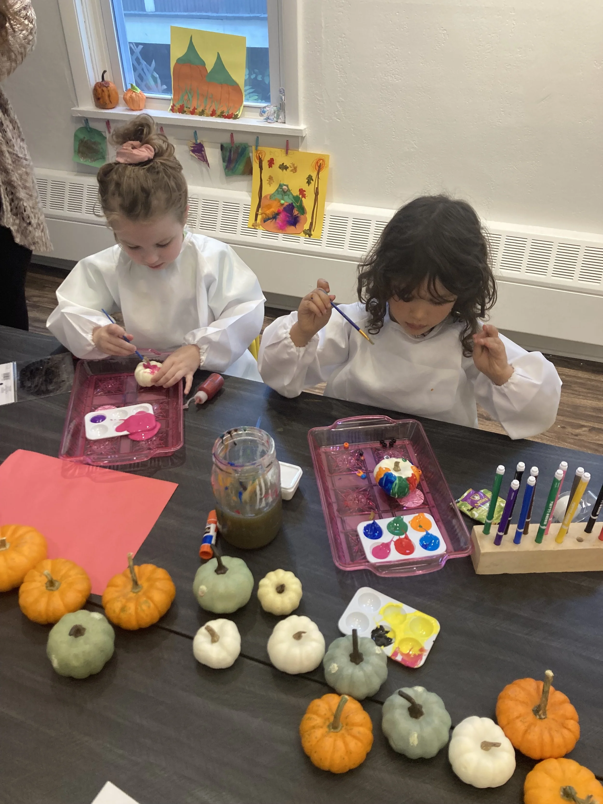 Two young children painting small pumpkins at a table, surrounded by decorative pumpkins, with artwork hanging on the wall behind them.