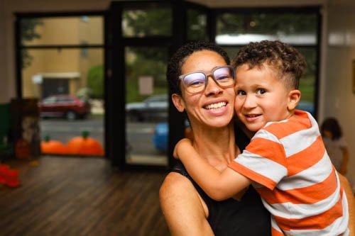 A woman wearing glasses holding a young boy with curly hair and a striped orange and white shirt inside a room with large windows.