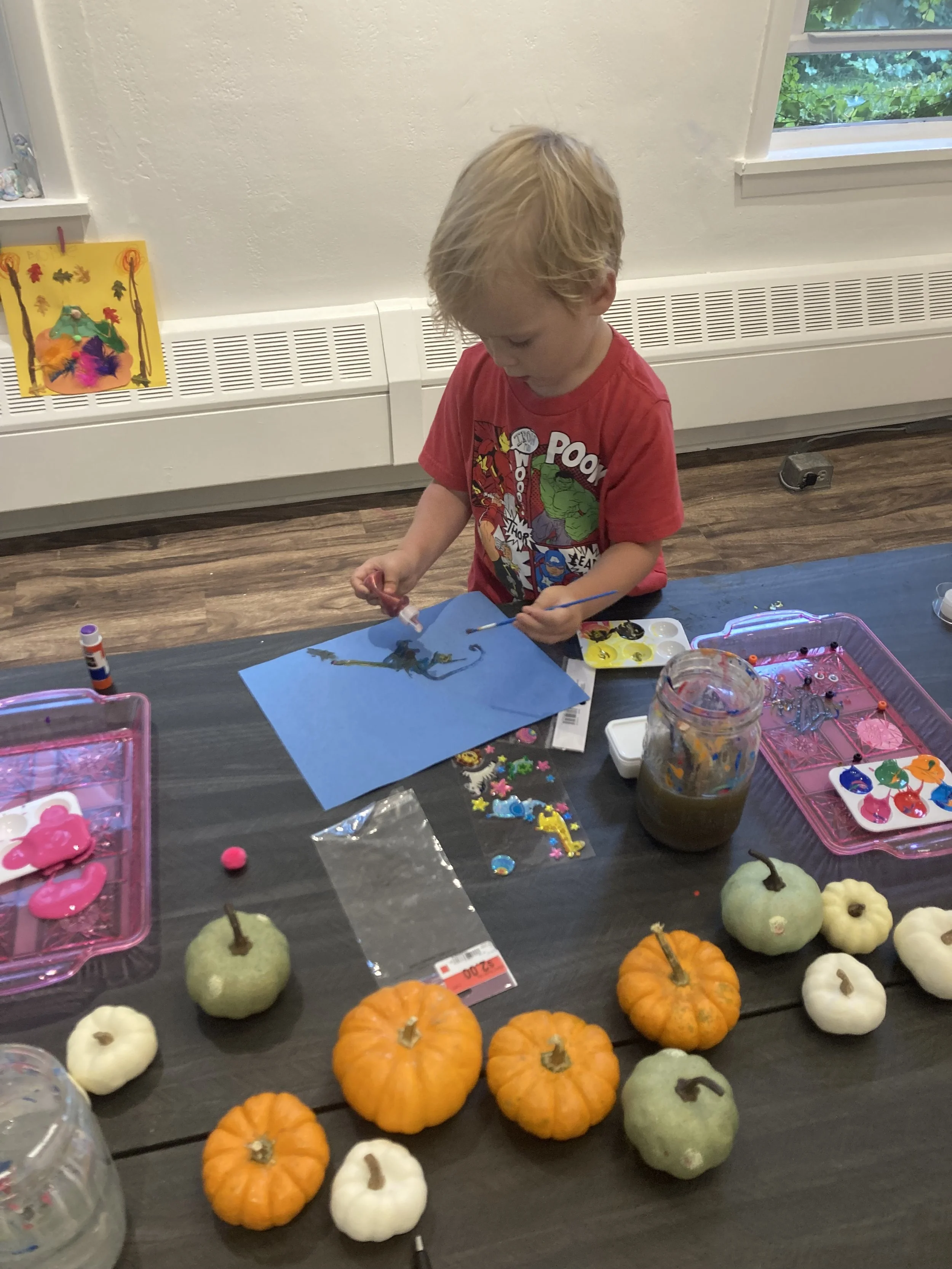 A young boy with blonde hair, wearing a red Marvel Avengers t-shirt, sits at a table doing arts and crafts. The table is decorated with pumpkins, craft supplies including glue, stickers, and paint, and a jar with paint or glue. A window with greenery