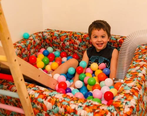 A young boy smiling and playing in a colorful ball pit with plastic balls inside a room.