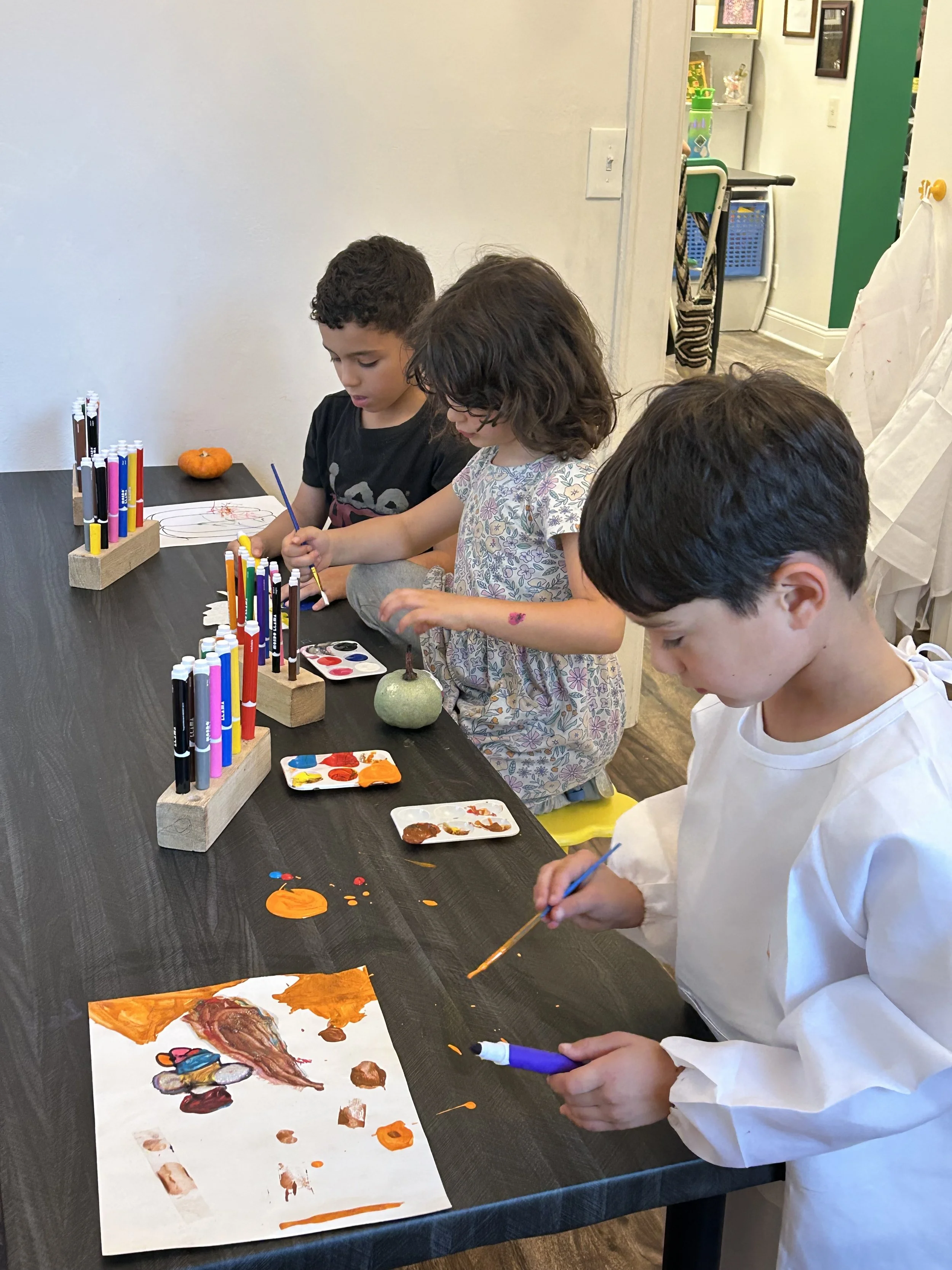 Four children sitting at a table, painting and drawing on paper with various art supplies in a classroom setting.