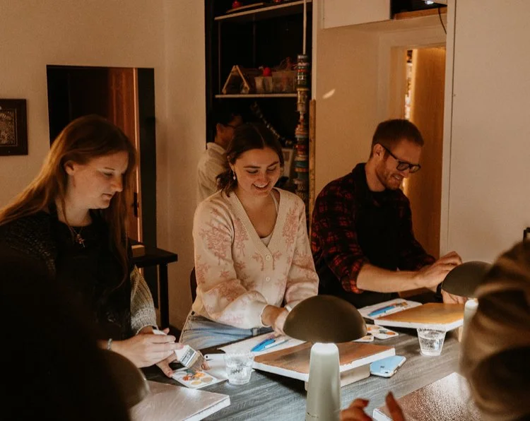 Four people sitting at a table, smiling and engaging with objects in front of them, in a cozy indoor setting.