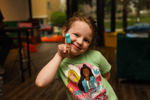 A young girl with curly hair smiling and holding a blue pill near her face inside a colorful room.
