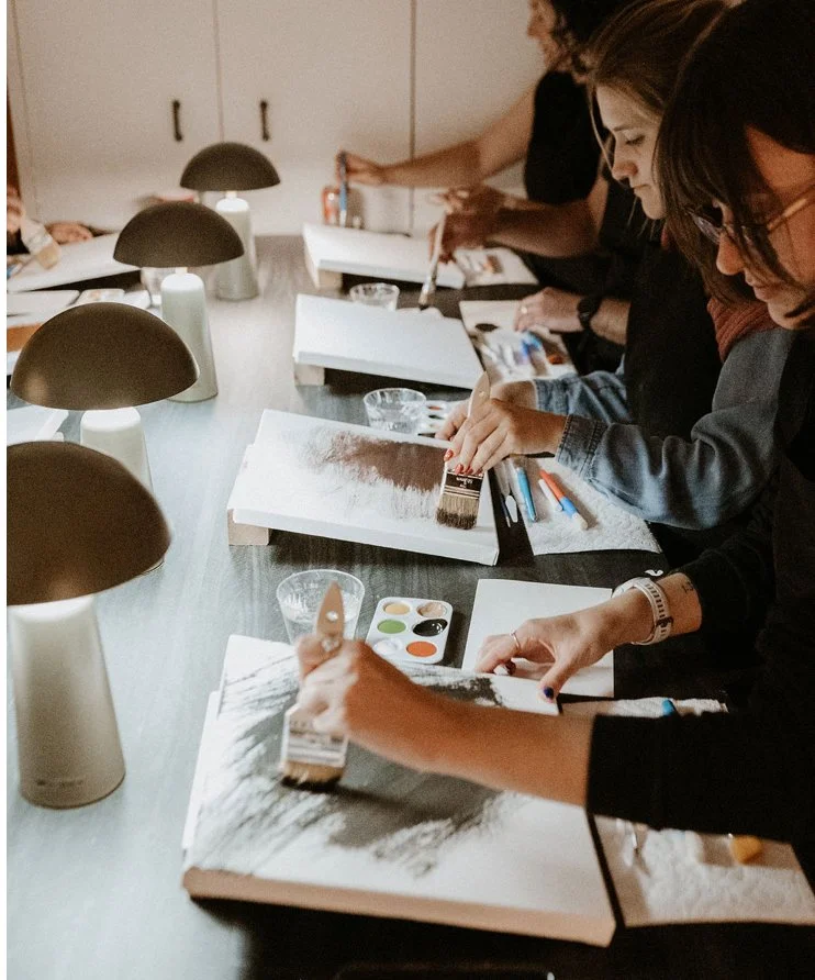 Group of people participating in an art workshop, painting black and white artworks on canvases with brushes and watercolor paints, illuminated by small table lamps.