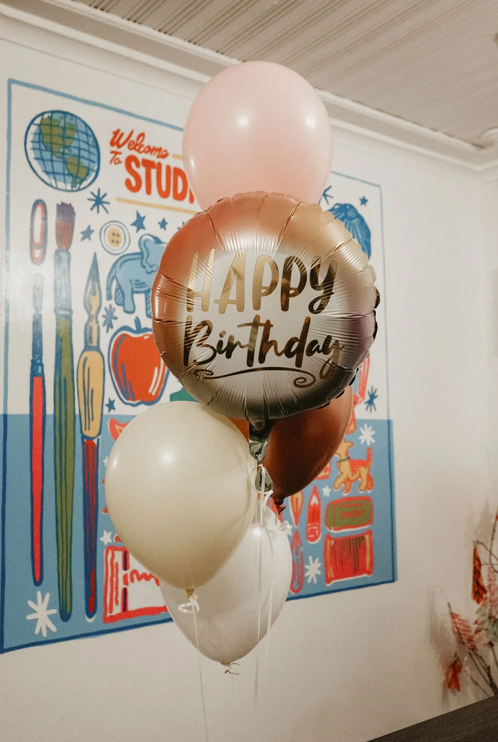 A cluster of balloons, including a round metallic balloon with 'Happy Birthday' written on it, pink, white, and brown balloons, in front of a colorful wall decoration with a classroom theme.