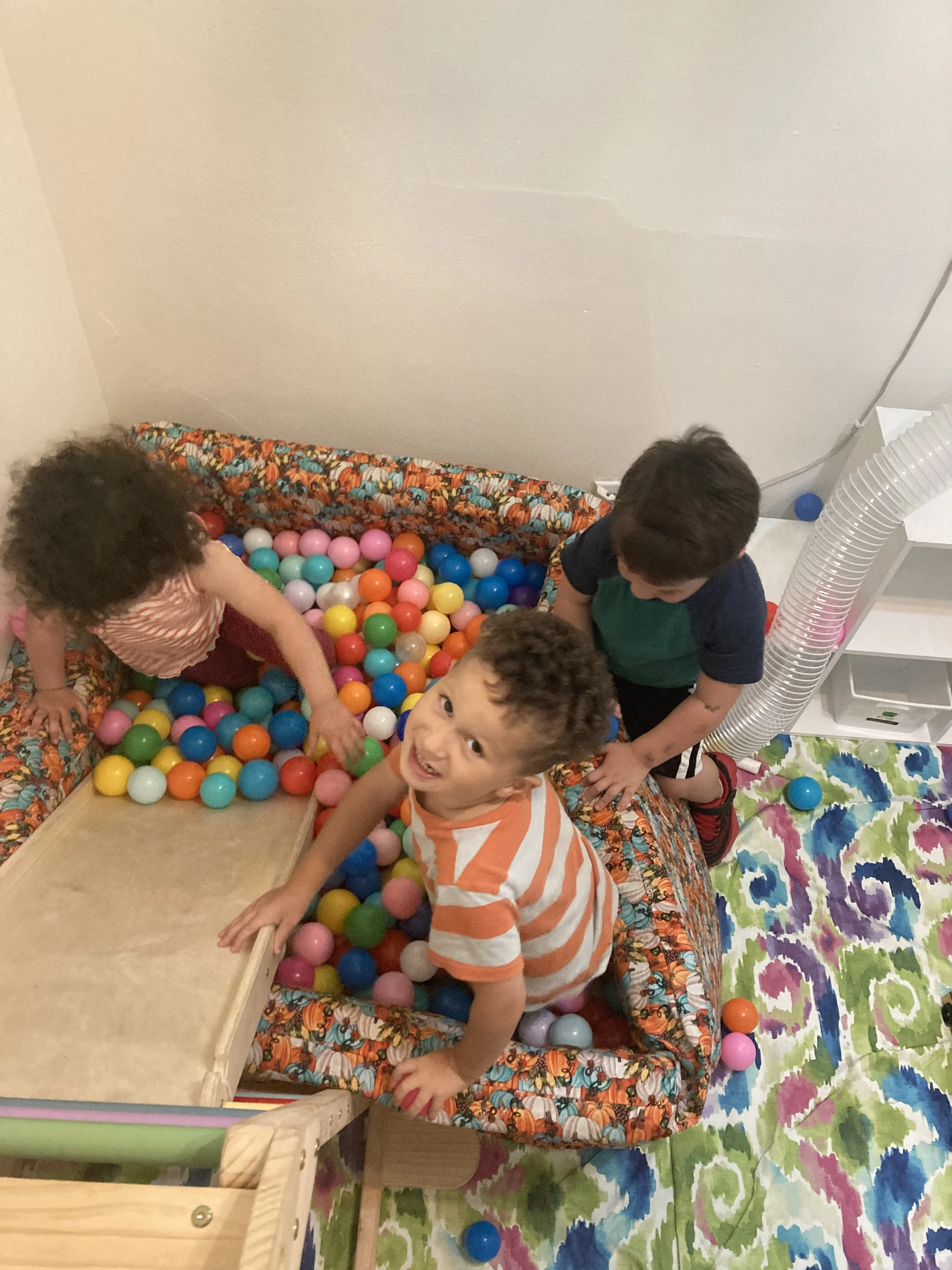 Three children playing in a ball pit with colorful plastic balls in a room with a painted carpet and a plain wall.