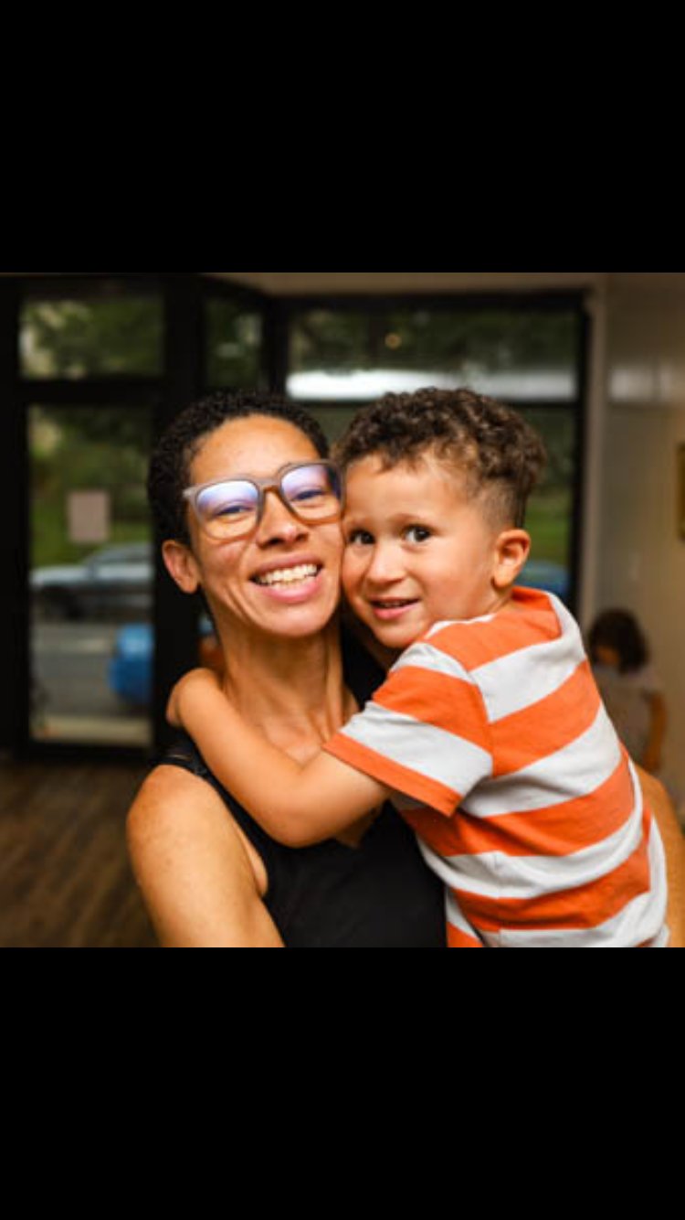 A woman with glasses and a young boy hugging smiling at the camera inside a building with large windows.