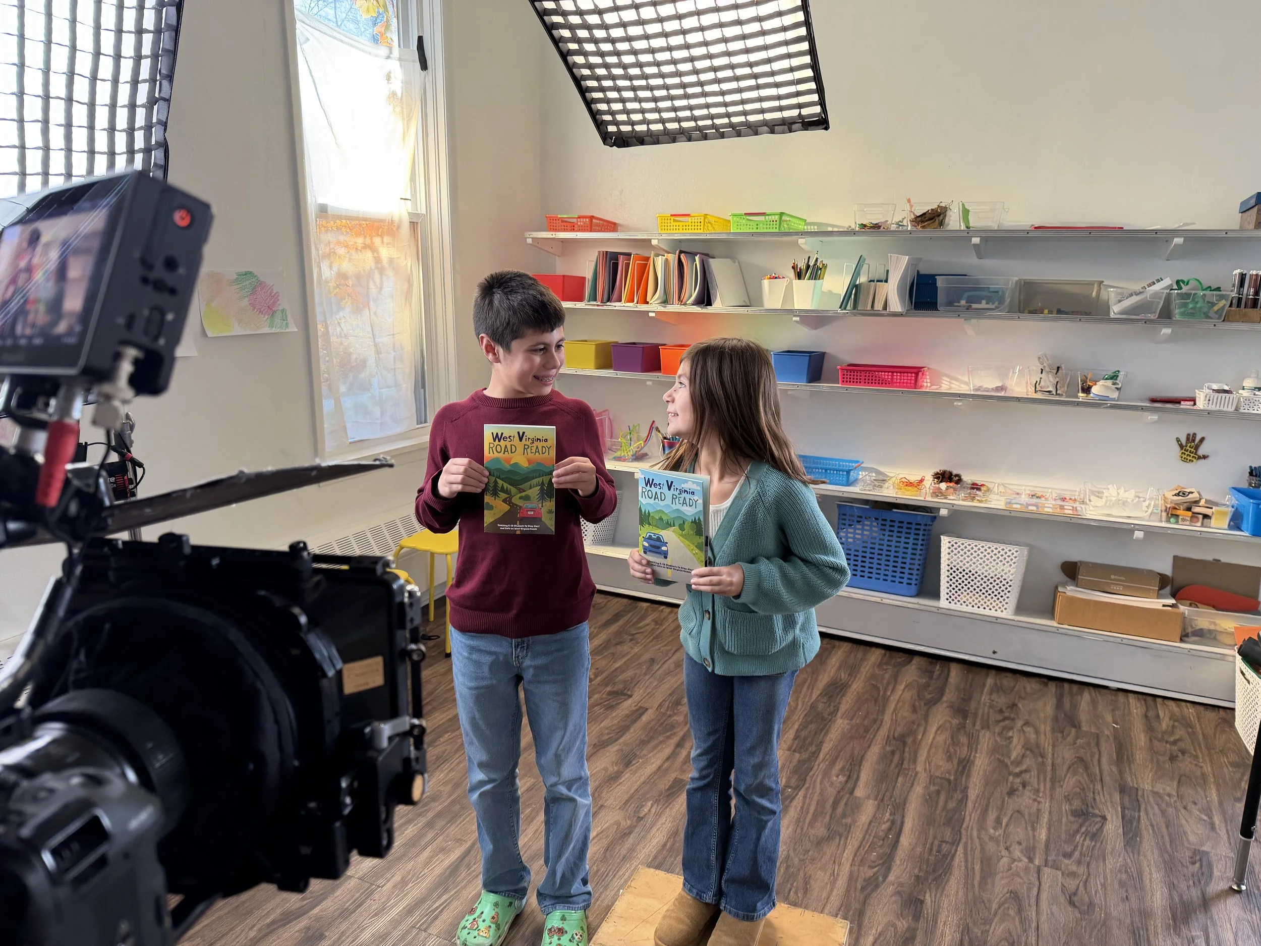 Two children standing in a classroom, smiling and holding books titled 'West Virginia ROAD READY,' with photography equipment in the foreground.