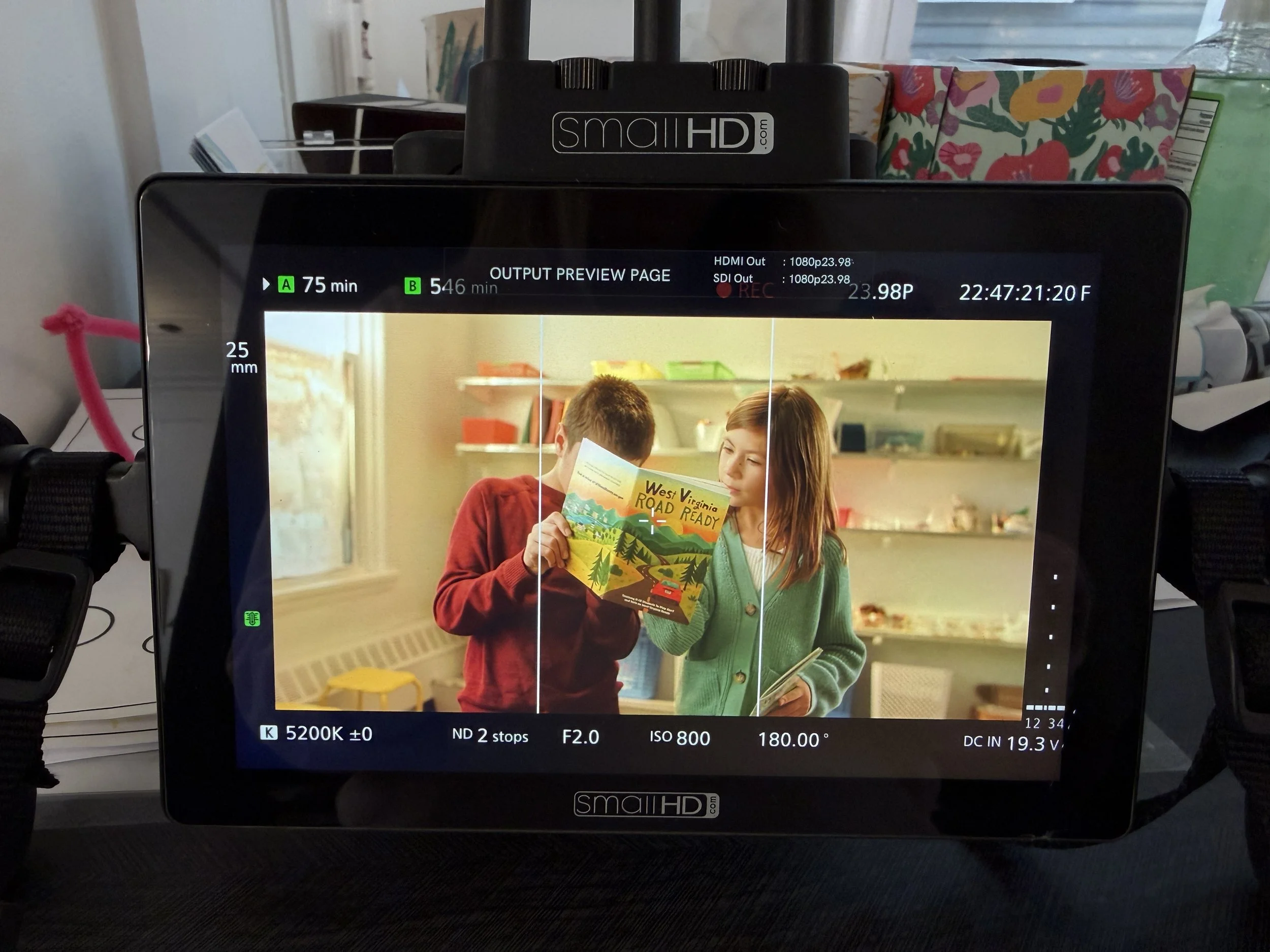 A monitor screen showing a photo of two children holding colorful books in a classroom. One child in a red sweater looks at the other girl in a green sweater, who is looking at the book she is holding.