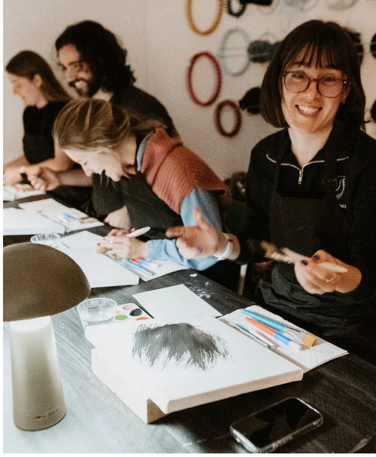 Group of smiling people sitting at a table engaged in an art activity, with art supplies and papers in front of them.