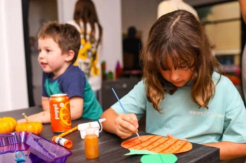 A girl painting a paper pumpkin at a table, with a boy smiling nearby, Halloween decorations, and crafts supplies on the table.