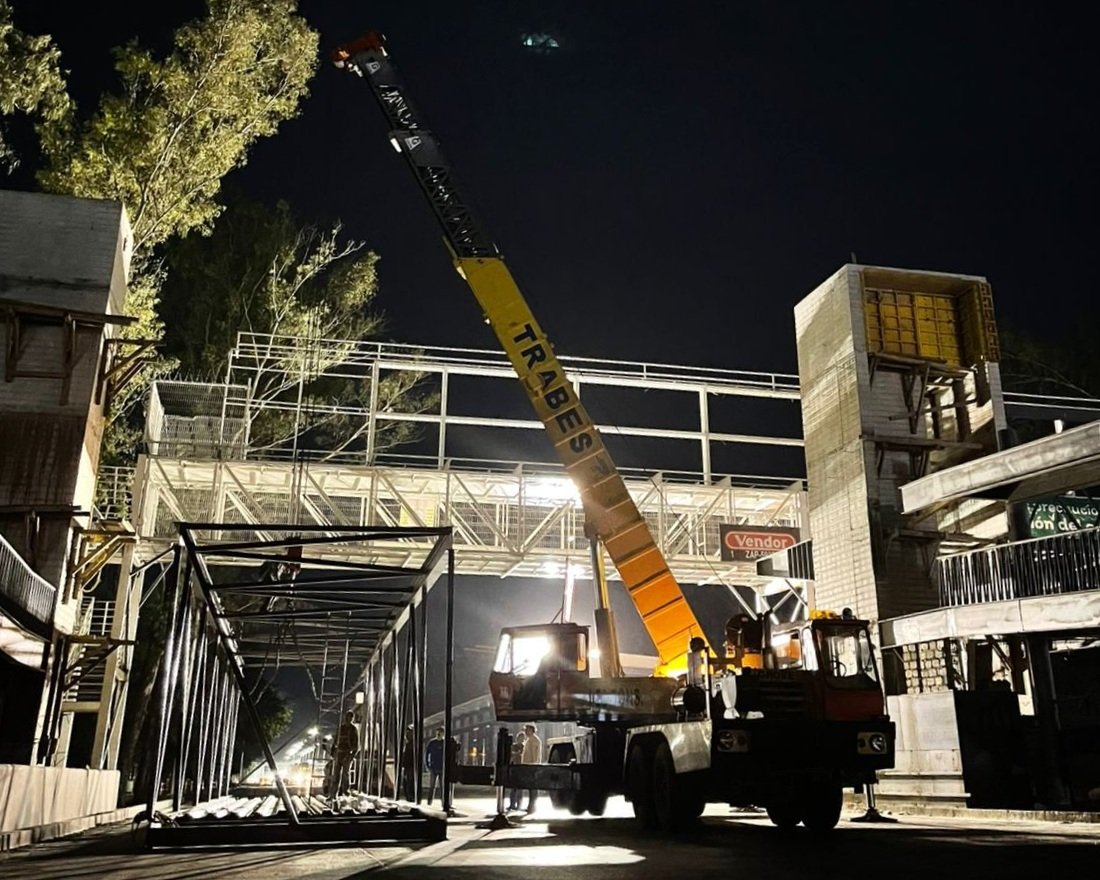 Night scene with a large crane lifting materials to a construction site, surrounded by unfinished buildings and a pedestrian bridge, illuminated by bright artificial lights.