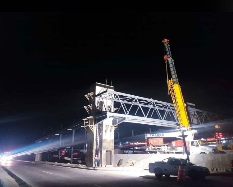 A construction crane is installing or repairing a bridge over a highway at night, with vehicles passing below and construction equipment visible nearby.