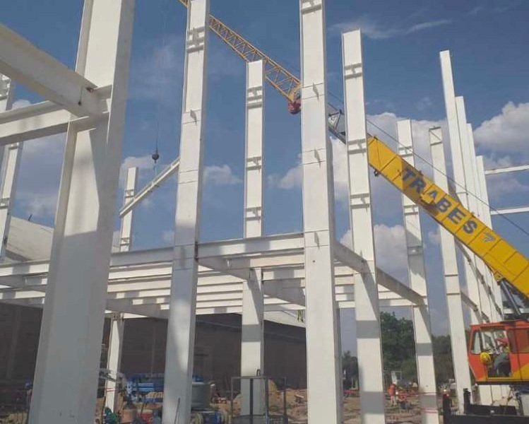 Construction site with steel beams being assembled, a yellow crane lifting materials, and a partially built structure.