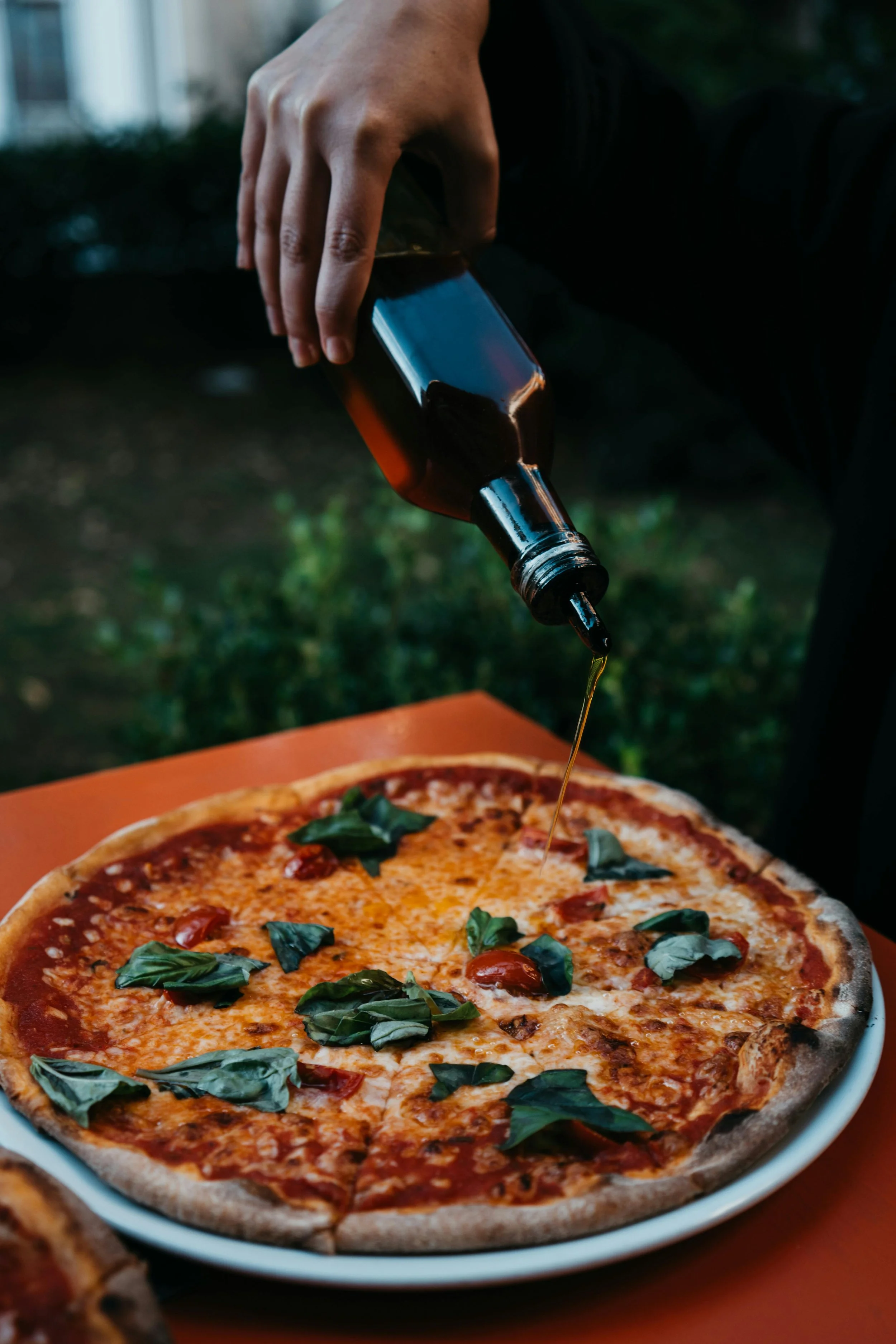 Person pouring infused olive oil onto a cheese and tomato pizza with basil leaves.