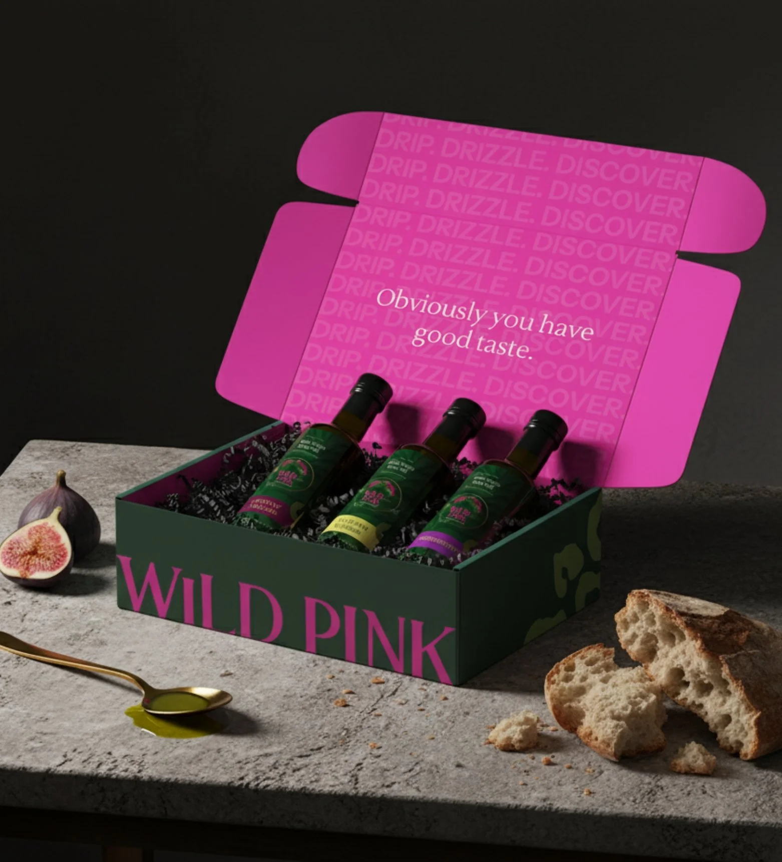 A box of three bottles of infused olive oil labeled 'Wild Pink' on a gray surface, with a halved fig, a spoon with infused olive oil, and torn bread nearby, and a pink card inside the box with the text 'Obviously you have good taste'.