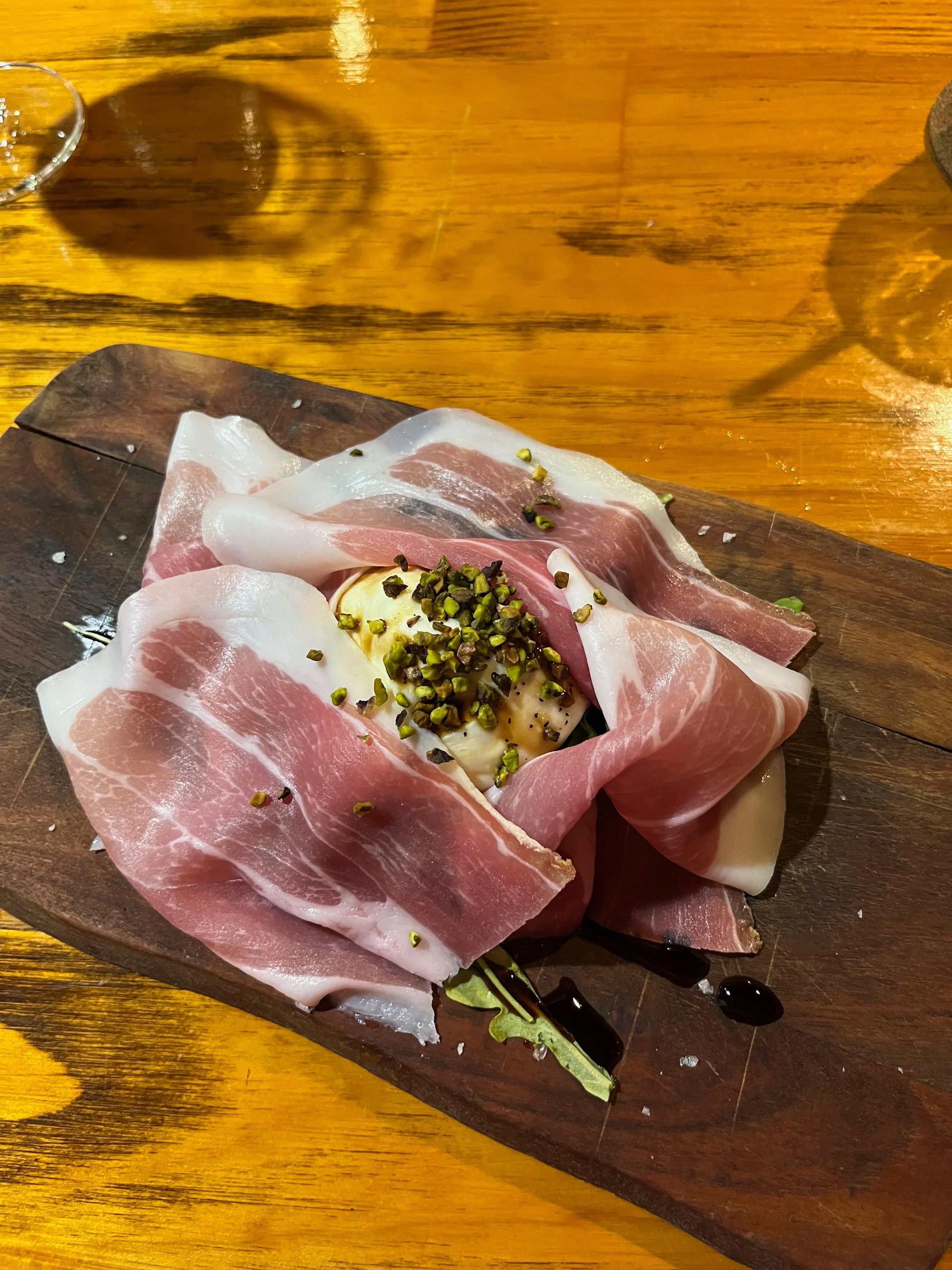 Sliced prosciutto with a dollop of soft cheese topped with chopped pistachios, served on a wooden platter with balsamic glaze.