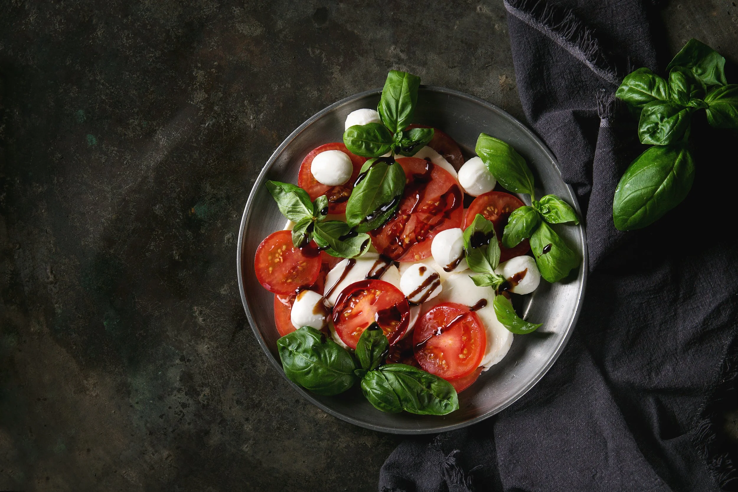 Caprese salad with sliced tomatoes, mozzarella balls, fresh basil leaves, balsamic glaze, and olive oil on a gray plate, placed on a dark textured surface with a black cloth.