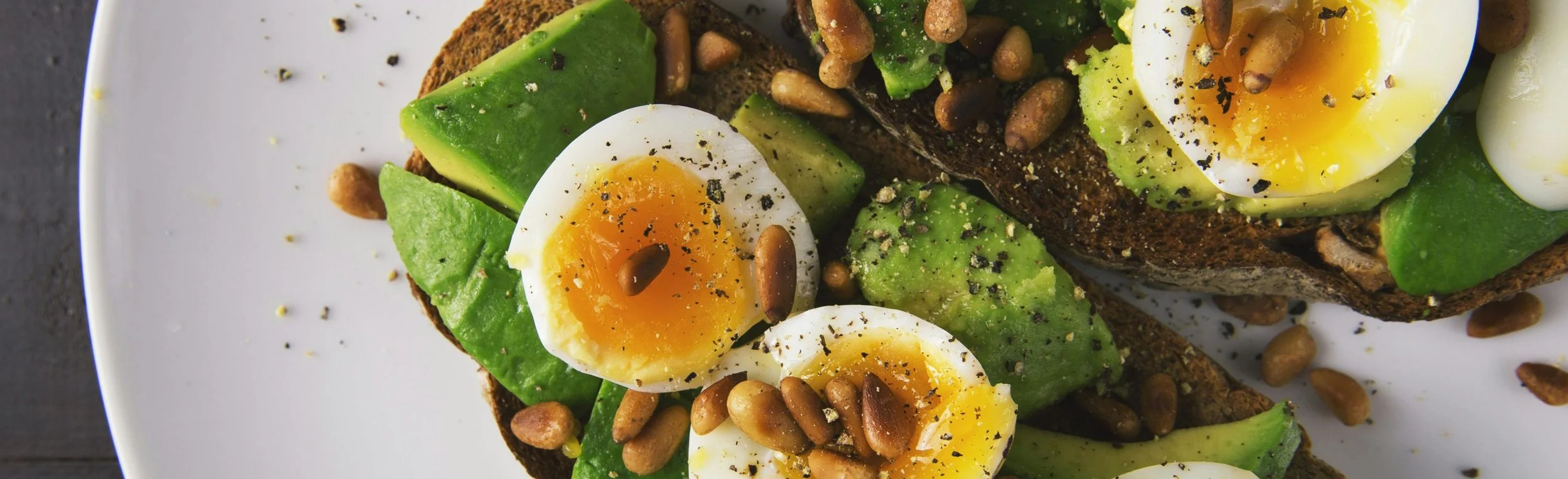 Open-faced avocado toast topped with soft-boiled eggs, pine nuts, black pepper, and salt on a dark slice of bread on a white plate.