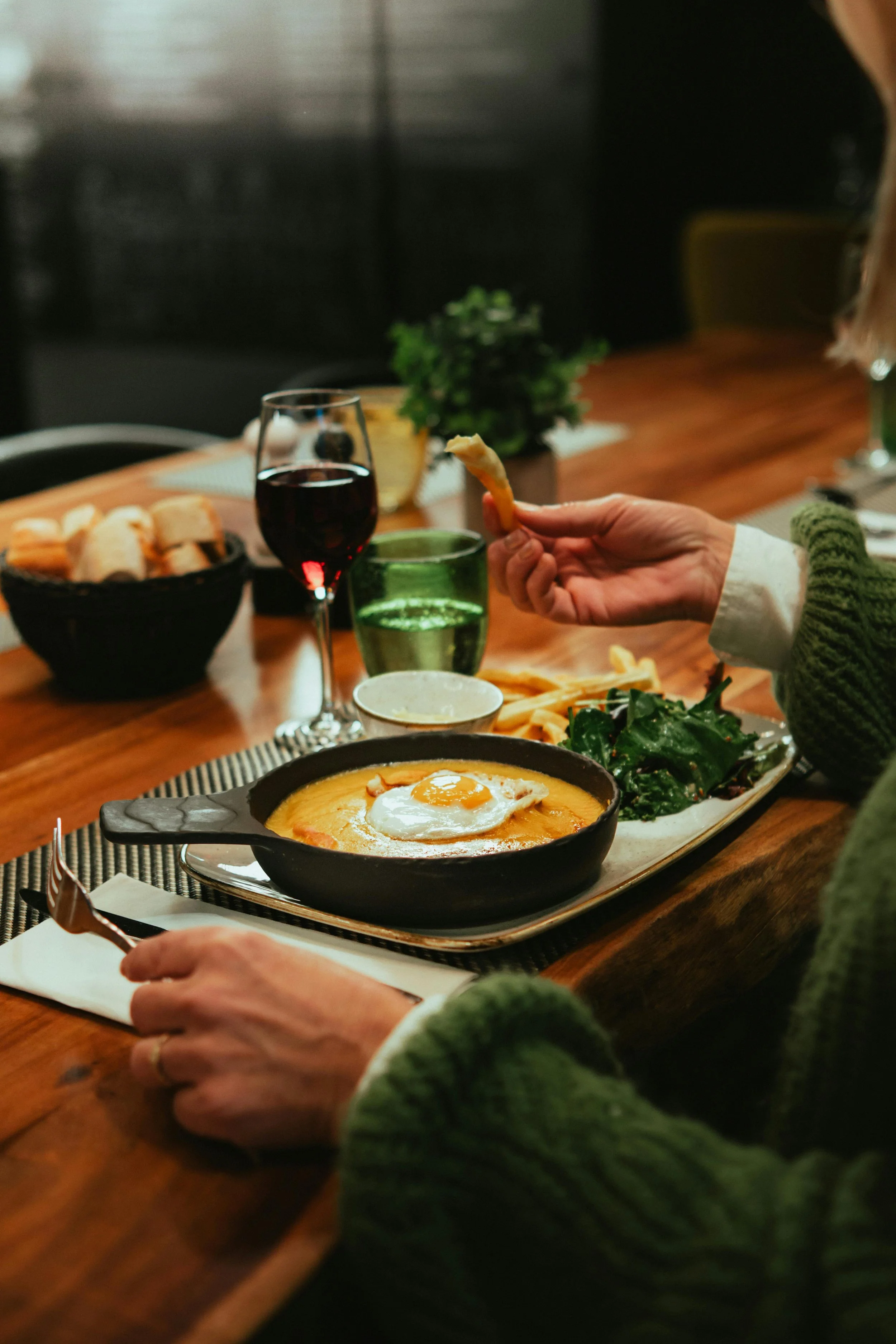 A person in a green sweater dining at a wooden table with a skillet containing baked eggs, accompanied by a side salad, with a glass of red wine, a glass of water, and bread in the background.