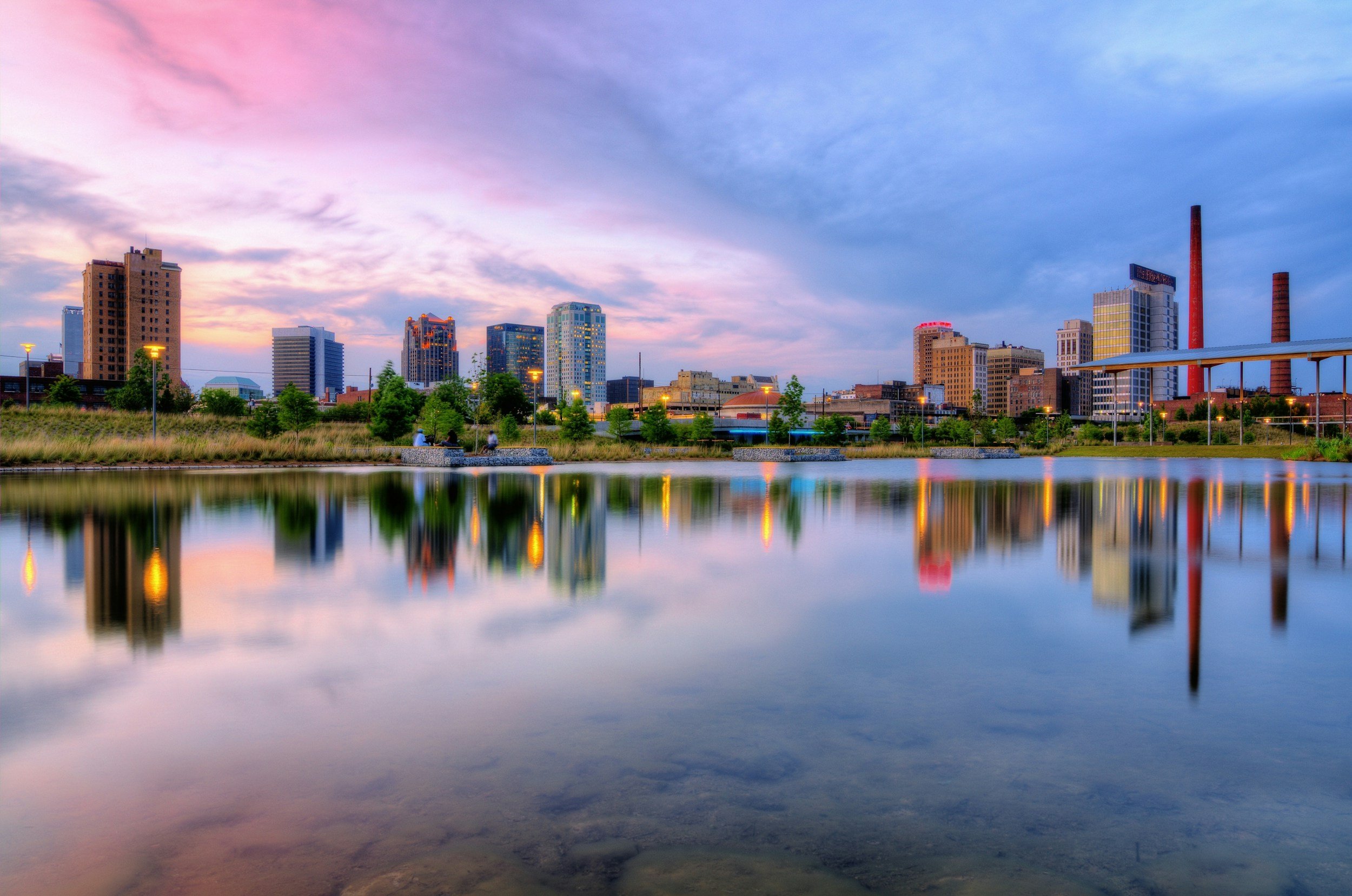 A panoramic view of a city skyline at sunset with tall skyscrapers, a harbor with boats, and urban parks with green trees.