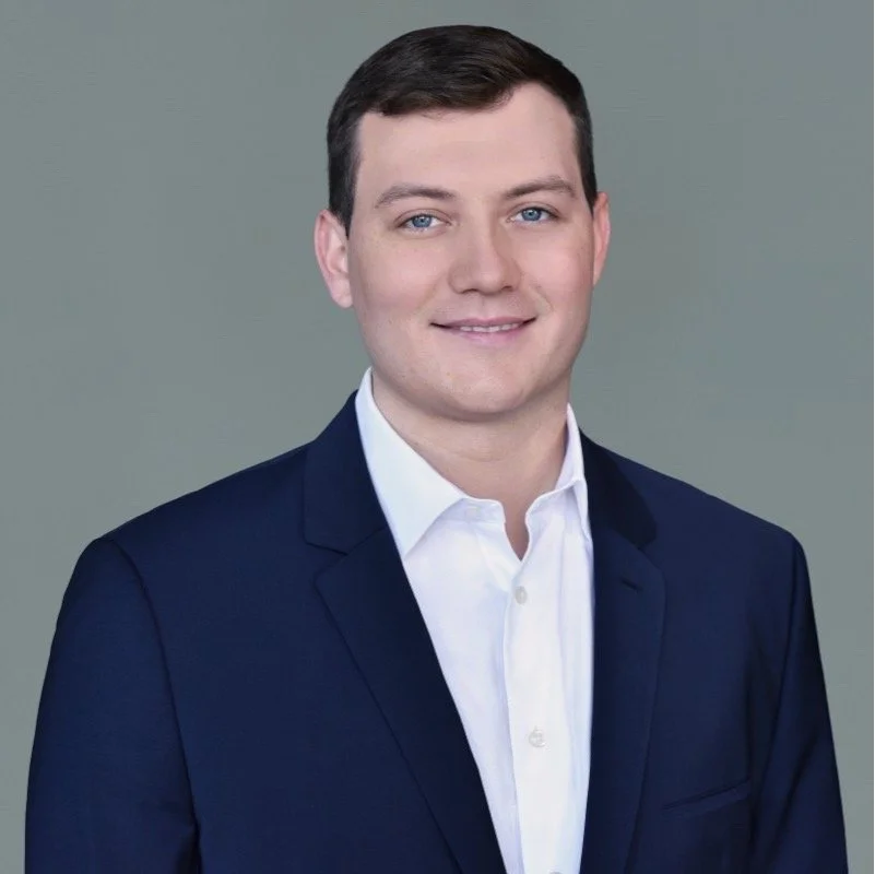 A young man with dark, curly hair and a bright smile, wearing a white button-up shirt with small black dots, standing outdoors in front of a building with brick and stairs, during daylight.