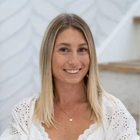 A young woman with long, wavy blonde hair, smiling, wearing a black jacket, standing outdoors with a cityscape and tall buildings in the background.