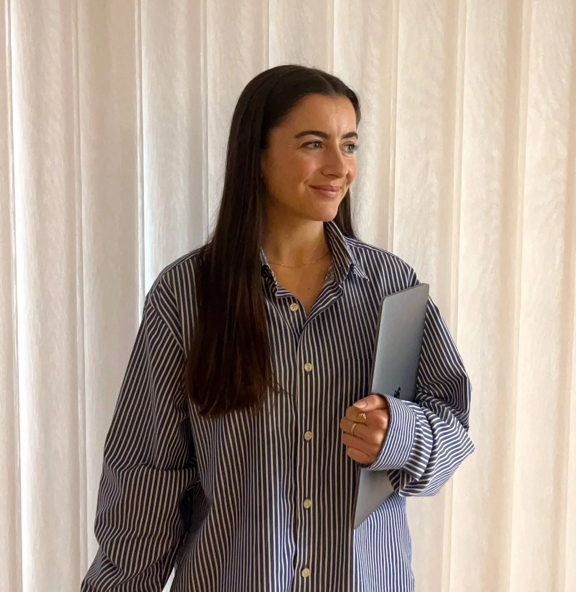 A woman with long dark hair wearing a striped shirt is standing in front of a white curtain, holding a gray tablet in her right hand and looking slightly to her right with a gentle smile.