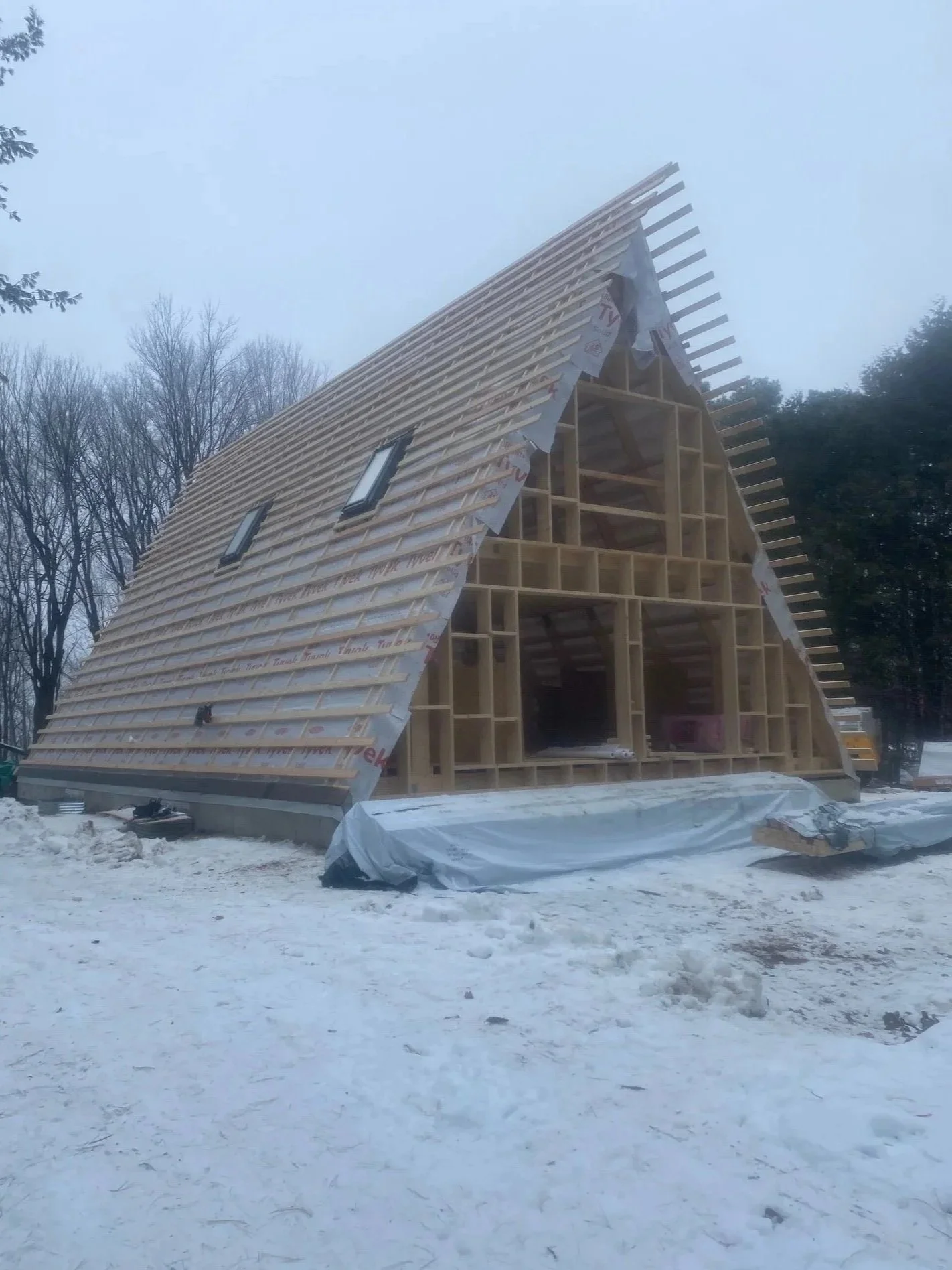 Building under construction with wooden framing and partially installed roof, snowy outdoor environment with leafless trees.