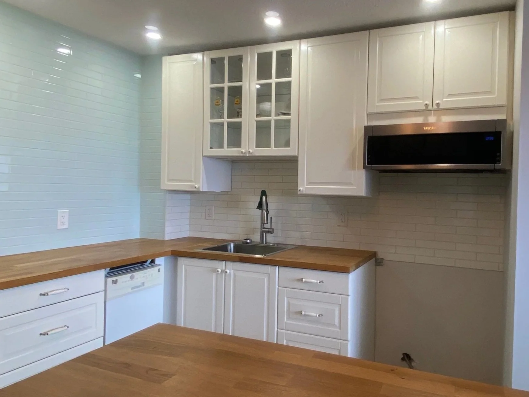 Kitchen with white cabinets, wooden countertops, a sink with a stainless steel faucet, a microwave oven, and tile backsplash. The wall on the left has light teal tile, and there is an area with outlets and a dishwasher below the countertop.