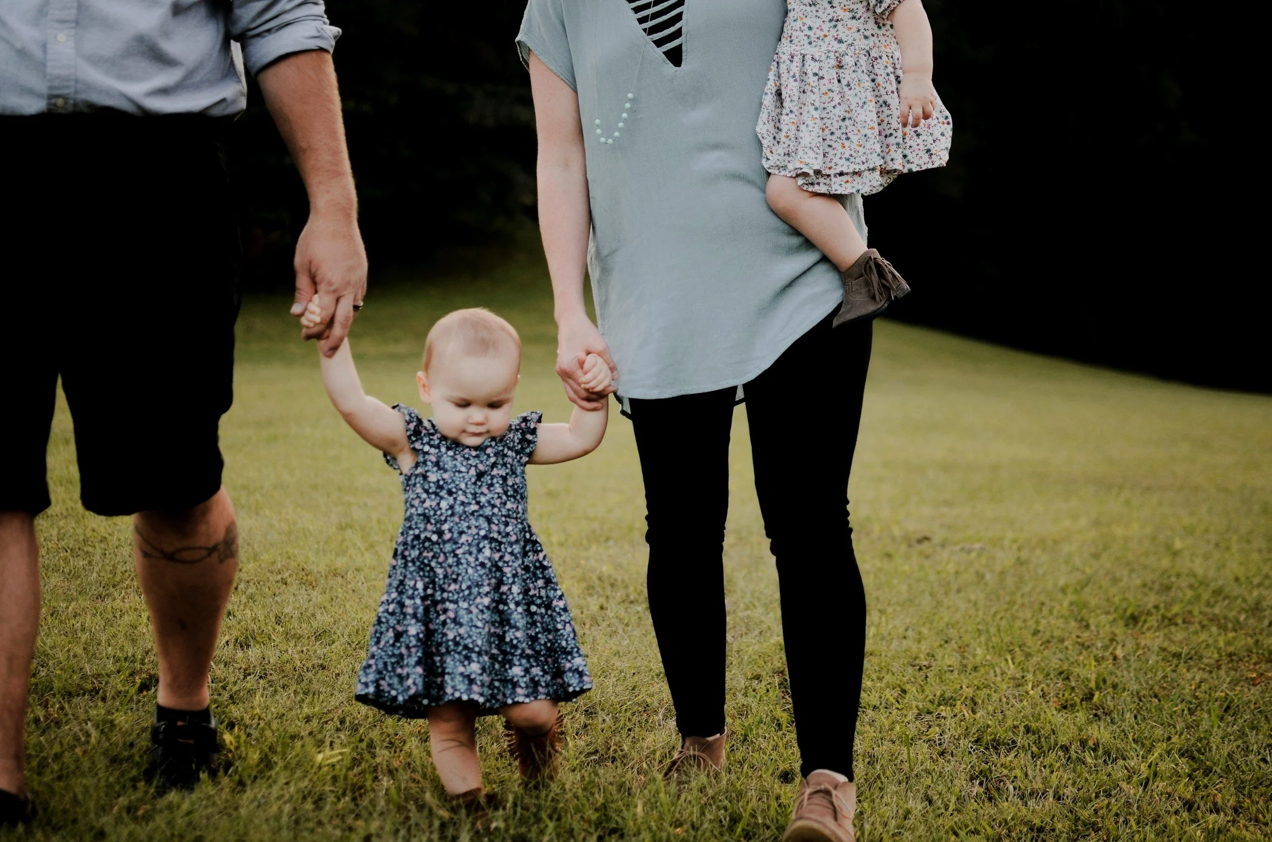 A family of three, including a man, a woman, and a toddler girl, holding hands and walking on a grassy field outdoors. The woman is carrying a young child on her hip.