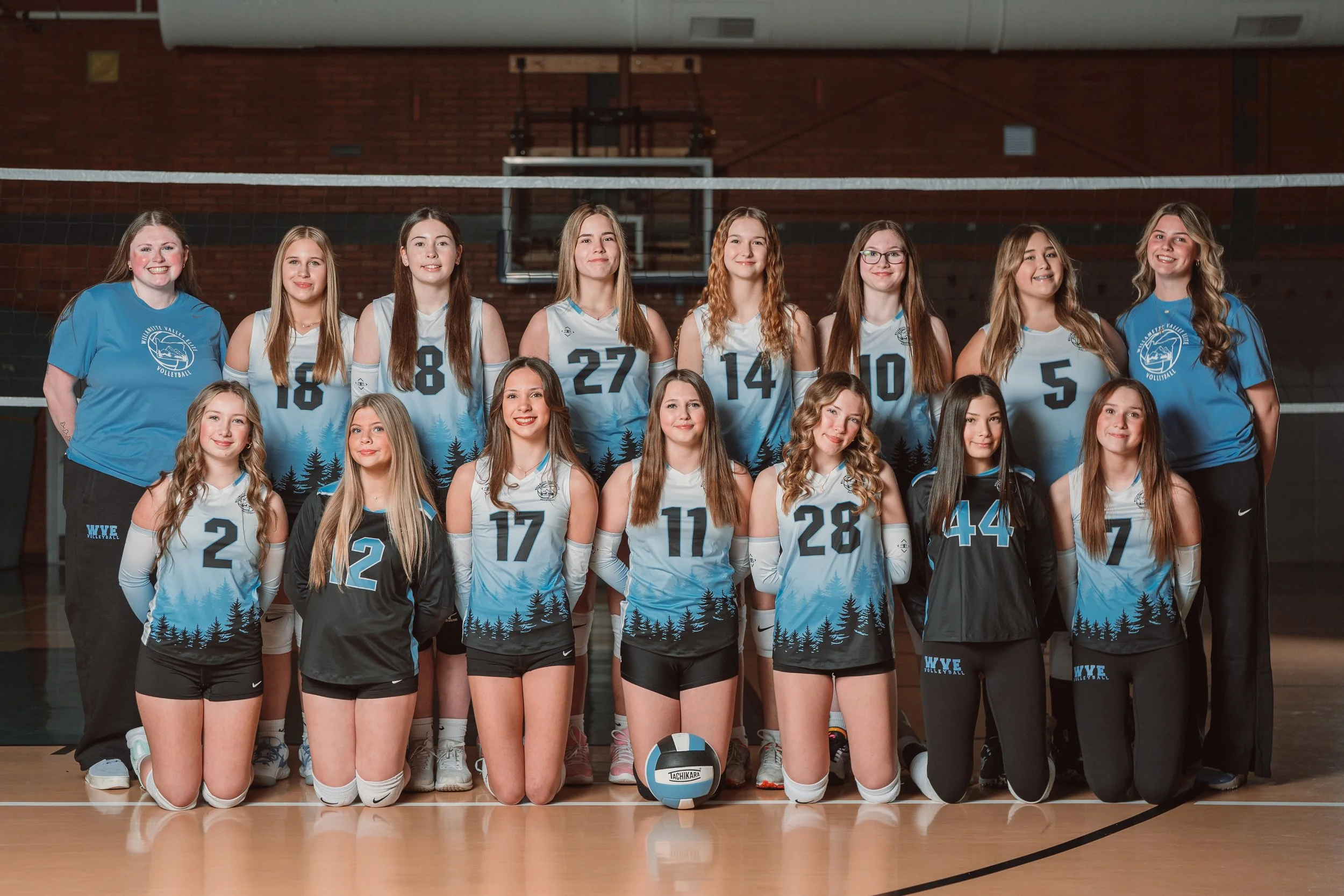 A girls volleyball team team posing for a photo in a gymnasium, with players wearing black jerseys with blue accents and numbers, and coaches on either end.