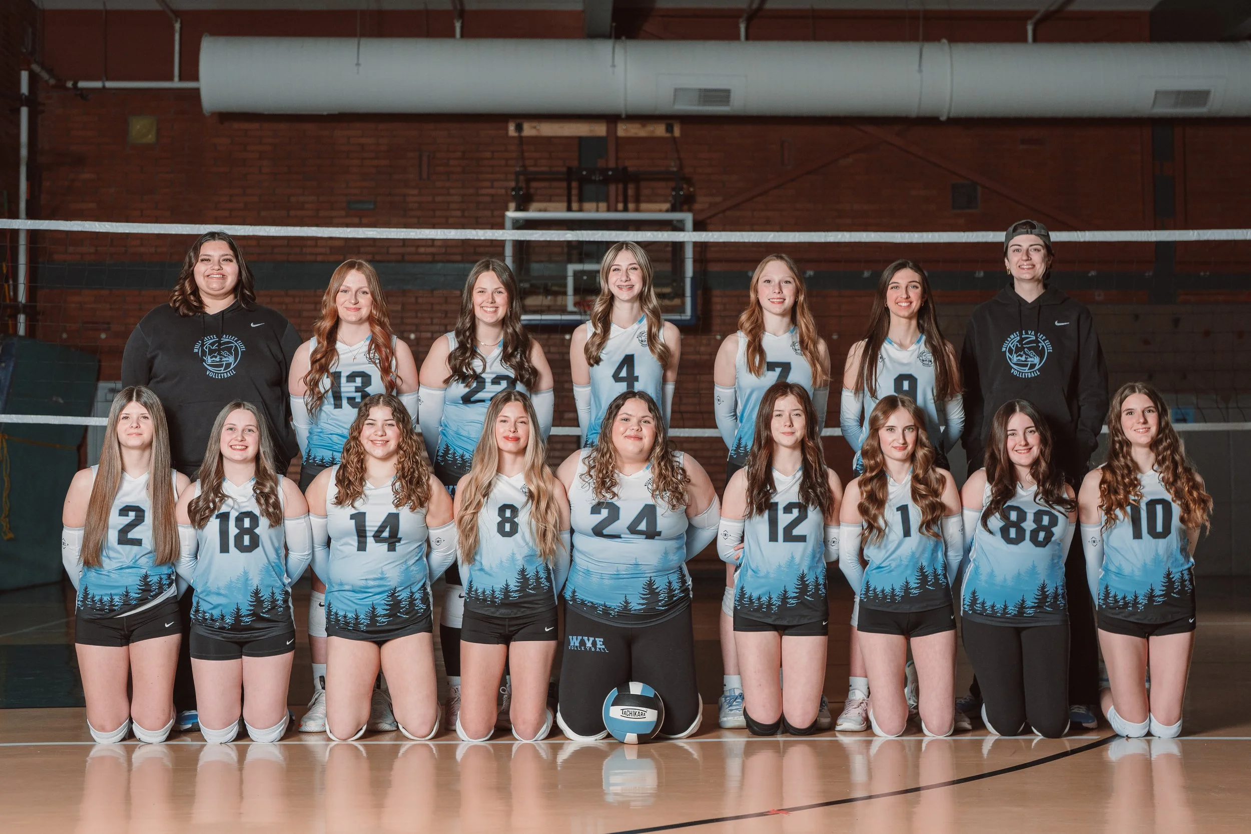 A volleyball team group photo indoors with players in black and blue jerseys and coaches wearing blue shirts.