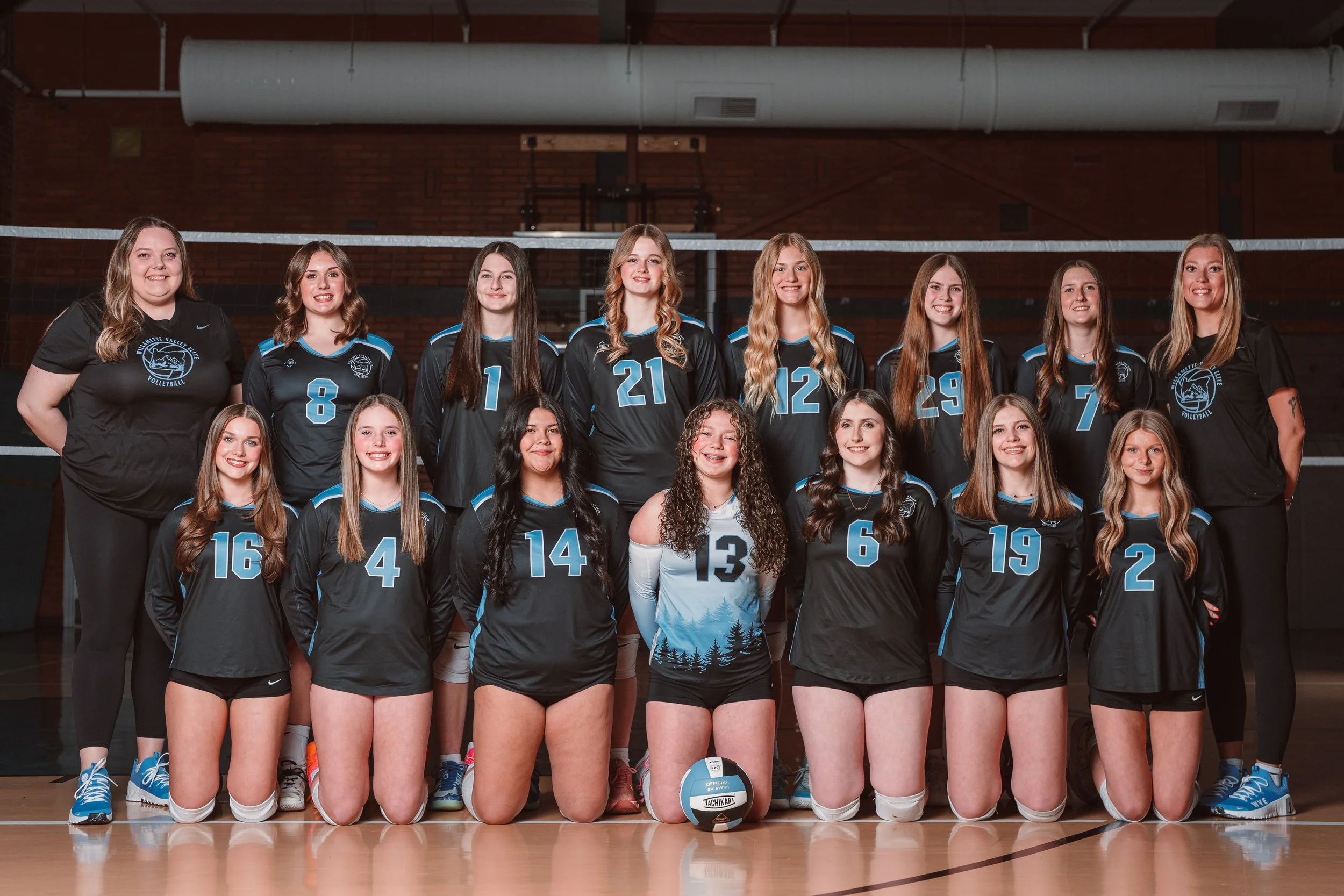 A girls' volleyball team posed for a team photo in a gymnasium. They are wearing light blue jerseys with white numbers and black shorts, with the exception of the player in the black jersey holding a volleyball. The background shows a volleyball net and a basketball hoop.
