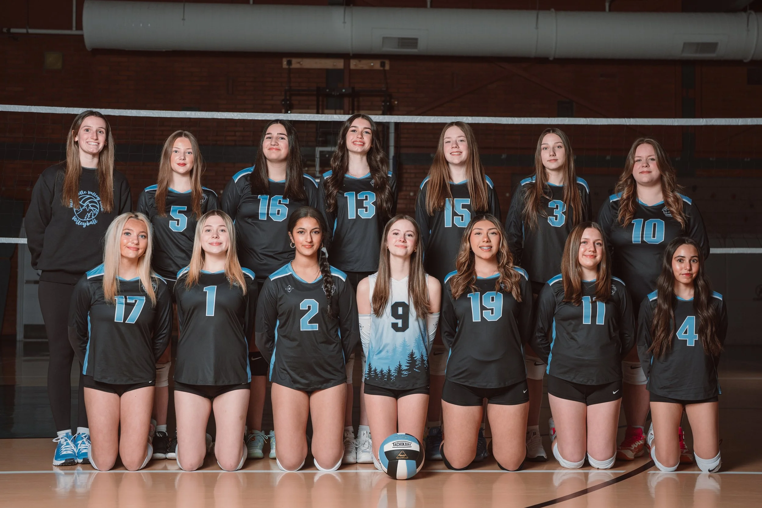 A girls volleyball team with 13 players in black and blue uniforms posing on an indoor court, with two coaches on either side. The players are standing and kneeling in two rows, in front of a volleyball net, with a brick wall in the background.