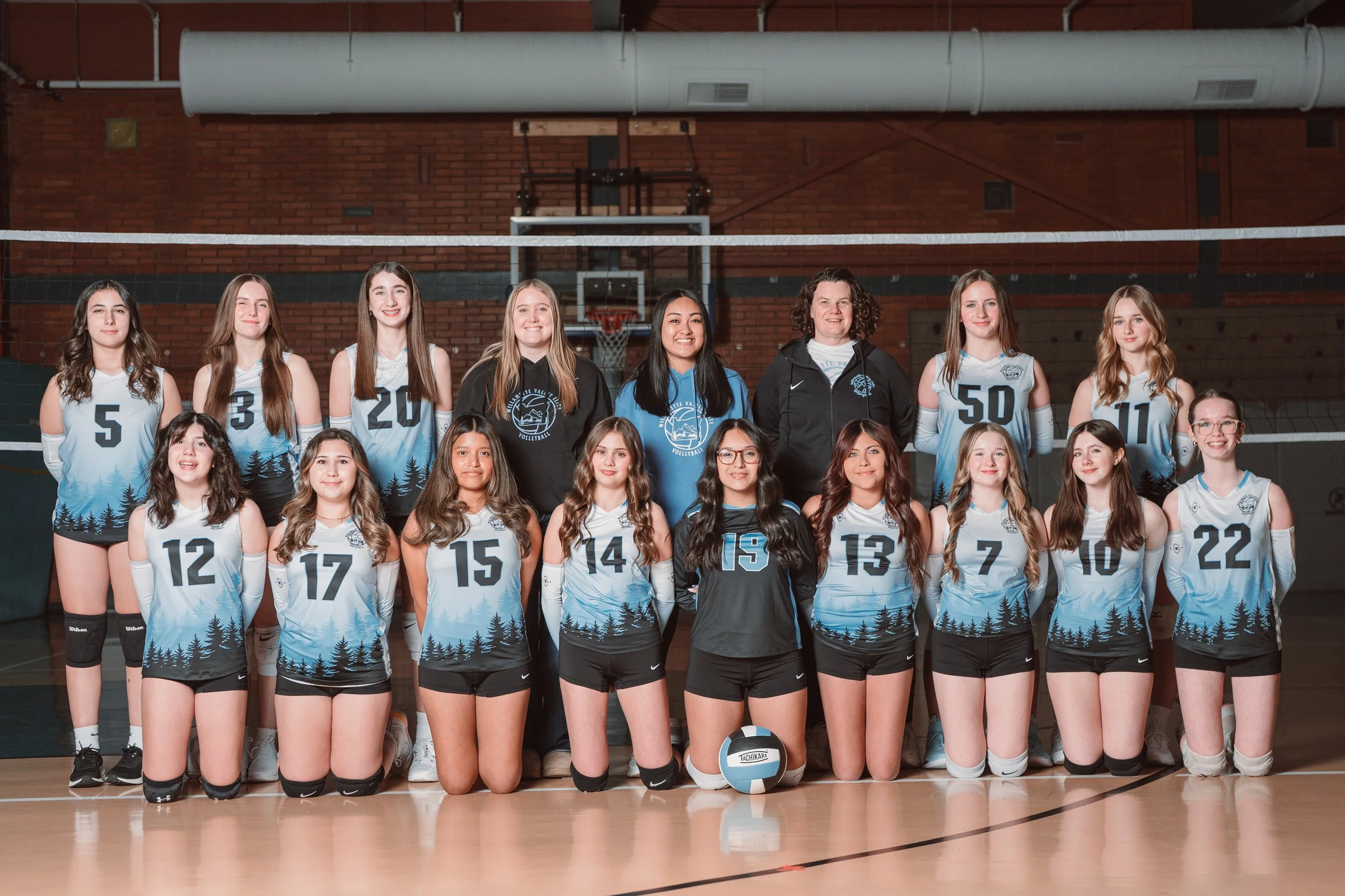 A girls' volleyball team posing for a team photo in a gymnasium, wearing black jerseys with blue accents and numbers.