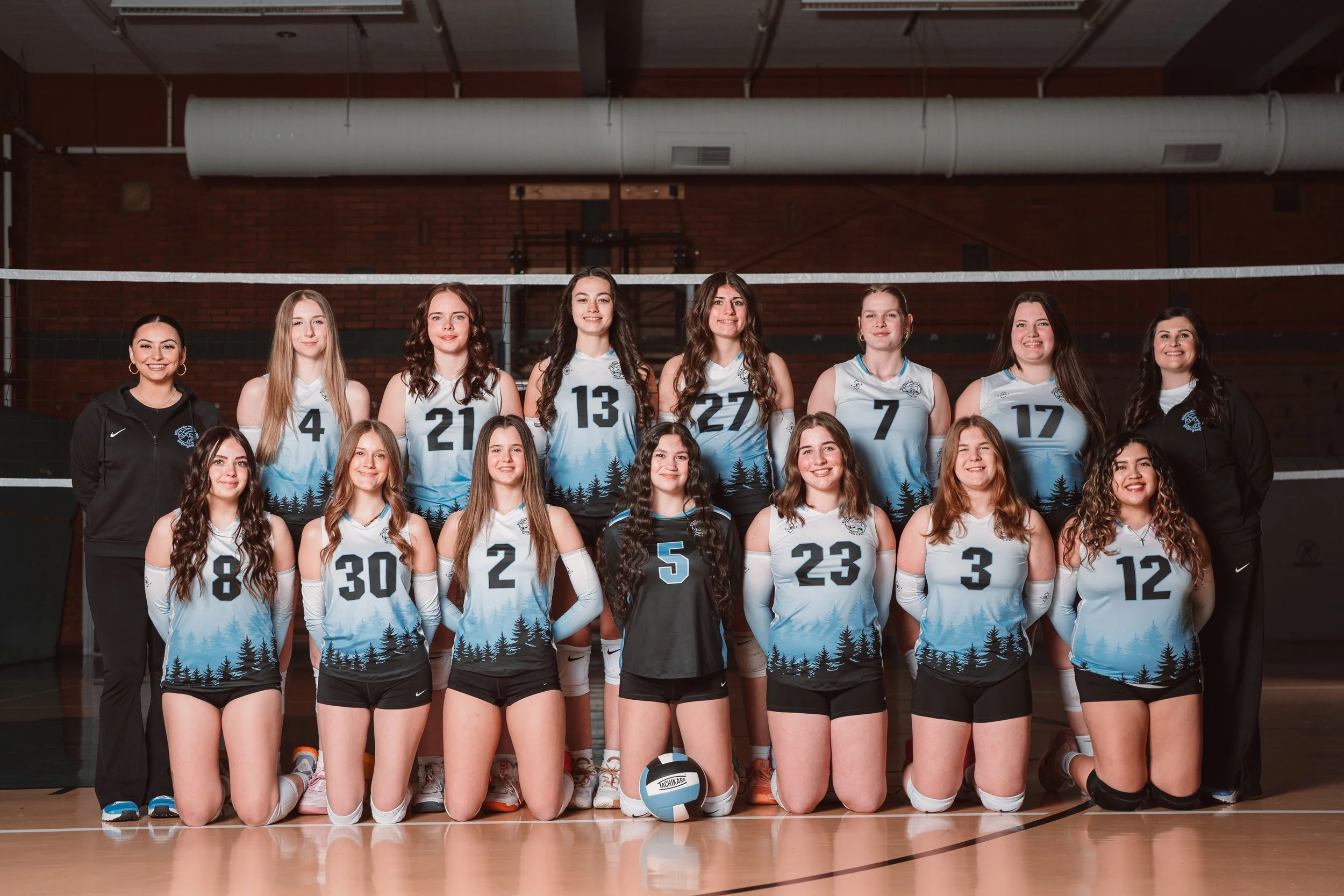 A group of young female volleyball players and their coaches posing for team photo in a gym, wearing blue jerseys and kneeling on the basketball court.