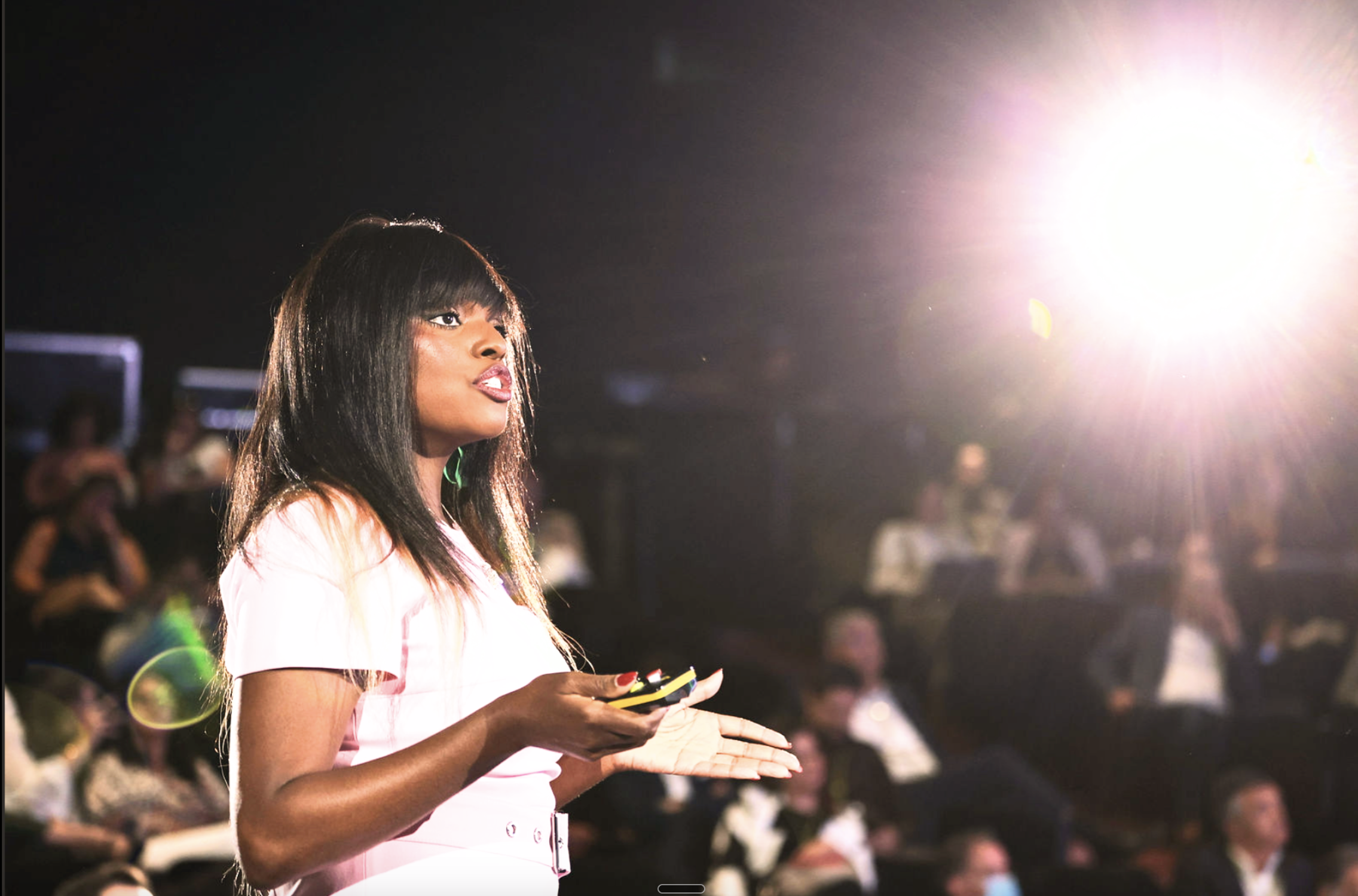 A woman with long dark hair and bangs, wearing a white dress, is standing in a dark room with a bright light source to her right. She appears to be holding a yellow-black object, possibly a phone or remote control, and is looking slightly upward with an open mouth. There are blurred people in the background.