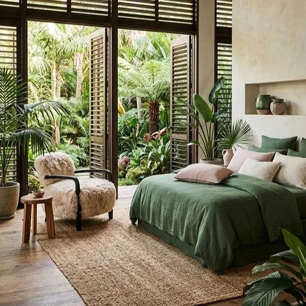 Bedroom with green bedding, beige pillows, and lush greenery outside the open balcony doors, decorated with plants and neutral decor.