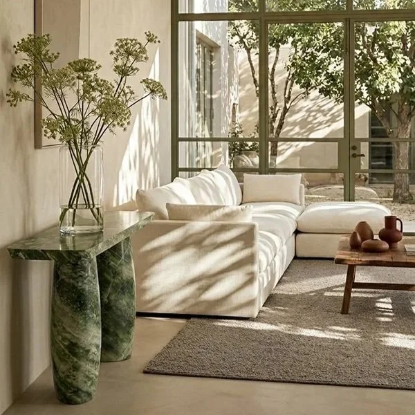 Sunlit living room with a white sofa, a wooden coffee table with decorative vases, a marble side table, a large window letting in natural light, and indoor plants casting shadows.