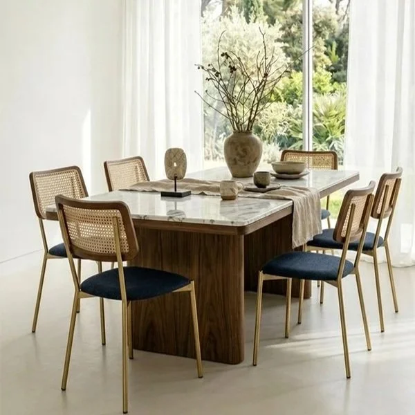 A modern dining room with a wooden table, six chairs with cane backs and dark cushions, a large vases with dried branches, and decorative stones, set near large windows with sheer white curtains showing greenery outside.