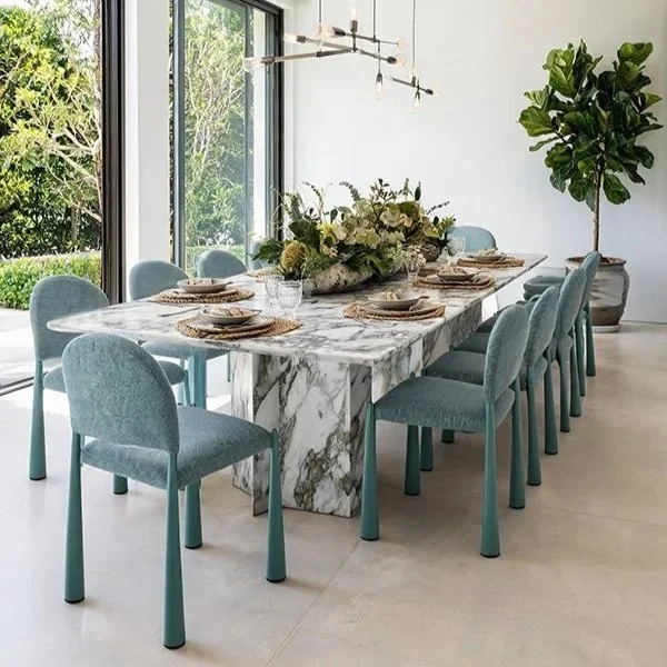 Modern dining room featuring a large marble table set with plates, glasses, and a floral centerpiece. Eight light blue upholstered chairs surround the table. Large windows with a view of greenery and a tall potted plant decorate the space.
