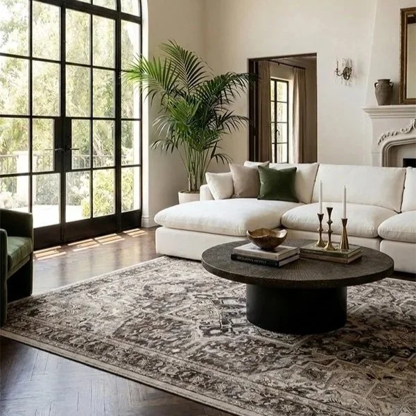 Living room with large window, white sofa, green cushions, a tall potted plant, fireplace, and a round coffee table with candles and books.