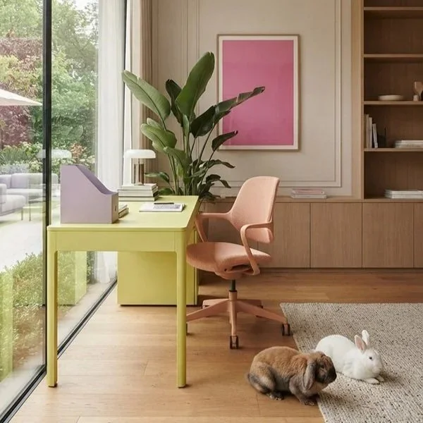 A home office with a yellow desk, pink office chair, green plant, and pink wall art, with two rabbits and a pug puppy on the floor.