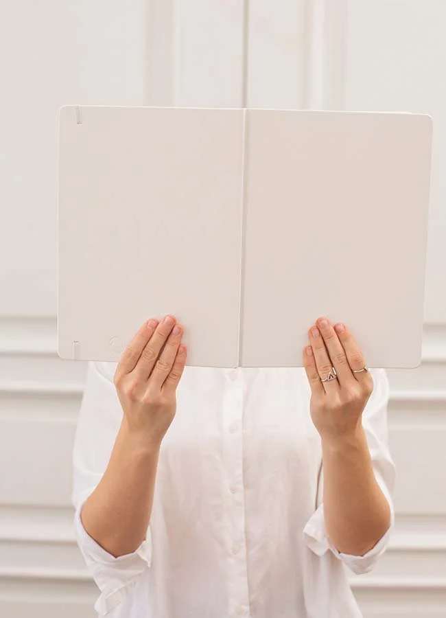 Person holding an open blank white notebook, obscuring their face, against a white paneled background.
